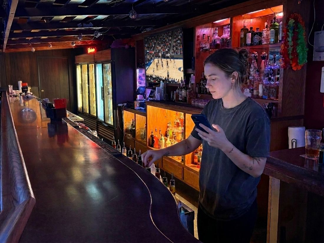 A bartender checking her phone behind a working bar, speed rail and back bar lit behind her.